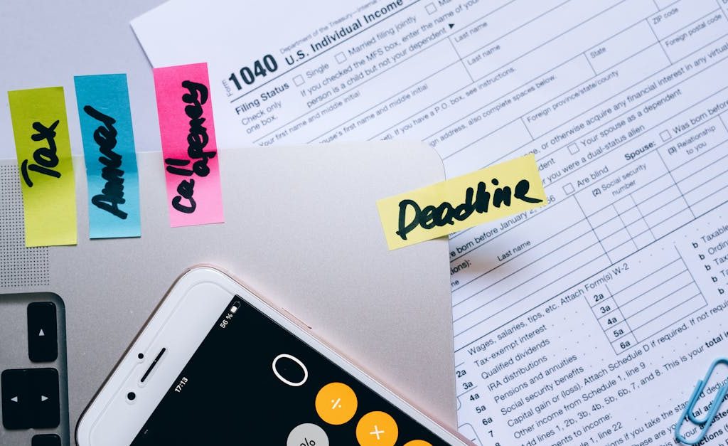 Office desk flat lay showing tax documents, calculator app on smartphone, sticky notes, and paperclips.
